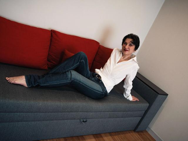 Maike75, pieds nus, portant une chemise blanche à boutons et un jean bleu, est assise sur un canapé gris avec des coussins rouges, le bras droit derrière elle, les jambes tendues..
