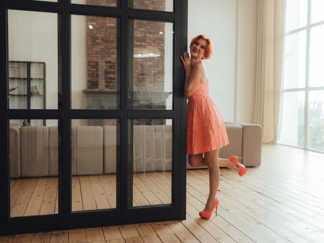 Maike75-hot with red hair, orange dress, and matching heels, stands playfully by a black divider, showing her ass, smiling, one leg raised on a wooden floor.