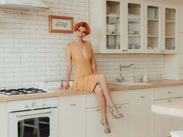 Maike75, capelli biondi, indossa un vestito giallo, seduta su un bancone bianco della cucina, gambe incrociate, mostra il sedere, con i tacchi alti, sorridente, con un muro di mattoni bianchi sullo sfondo..
