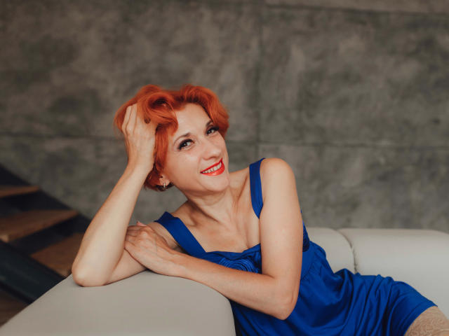 Maike75: capelli rossi, vestito blu e rossetto rosso, sorridente, appoggiata a un divano bianco, con una mano tra i capelli, che mostra leggermente il seno, in una posa rilassata e sensuale..