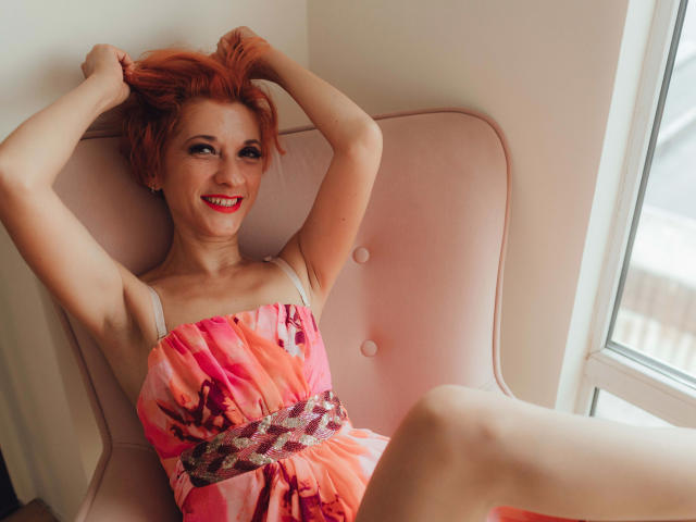 Maike75-atraente, com cabelo ruivo, sorrindo, vestindo um vestido floral rosa, com as mãos no cabelo, sentada em uma cadeira rosa, pernas abertas, batom vermelho, janela à direita..