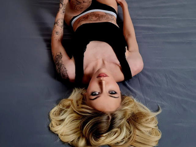Ginacolada, liegt auf dem Rücken, schwarzes Oberteil, weiße Unterwäsche, blondes Haar, Tattoos an Armen und Brust, graues Bett, blickt nach oben, Augen auf die Kamera gerichtet, Brüste sichtbar, Arme an den Seiten..