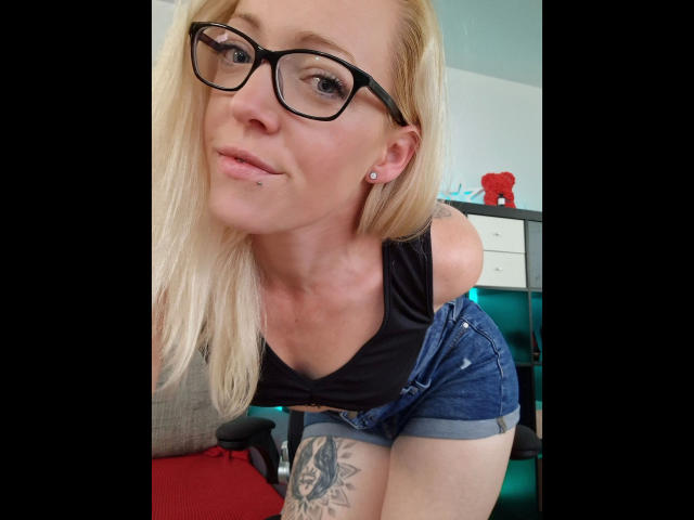 LeaKirsch – attraktiv, mit Brille, blonden Haaren, Nasenpiercing, schwarzem Oberteil, Jeansshorts, Tattoo am rechten Oberschenkel, leicht nach vorne gebeugt, lächelnd..