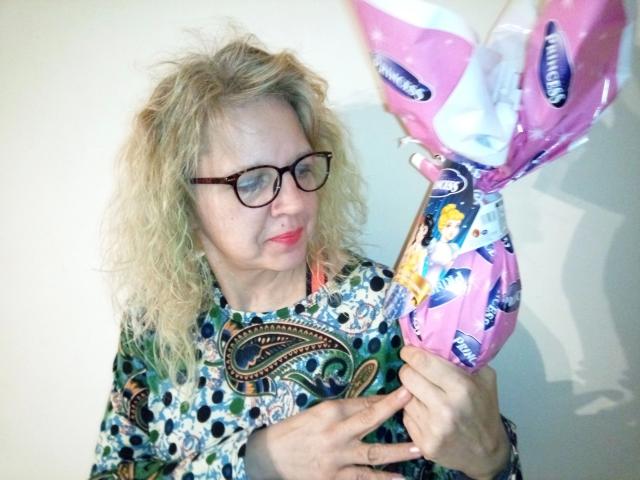 CuteDebby – attraktiv mit lockigem, blondem Haar und Brille, trägt ein farbenfrohes, gemustertes Hemd, hält eine pinke Geschenktüte mit blauen und weißen Schleifen und blickt mit einem leichten Lächeln nach unten..