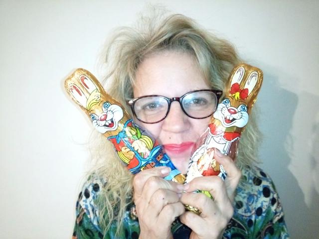 CuteDebby, bellissima con i capelli biondi spettinati, gli occhiali e il rossetto rosso, tiene davanti al viso due orecchie a forma di coniglio colorate, simili a caramelle, e indossa una camicia a fantasia..