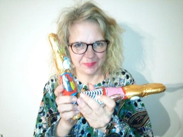 CuteDebby - aantrekkelijk met krullend blond haar, een bril en een kleurrijk shirt met een patroon, die twee belachelijke, in goudfolie verpakte, dildo-achtige speeltjes voor zich houdt en er lichtjes om lacht..