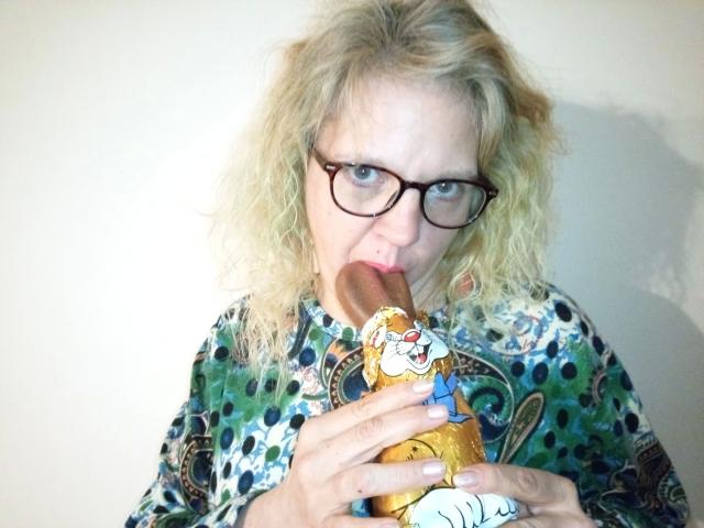 CuteDebby – hübsch, mit lockigem, blondem Haar und Brille, isst einen Hotdog mit einer cartoonartigen Verpackung, wobei ihr Mund um die Wurst ist, und trägt ein farbenfrohes, gemustertes Hemd..