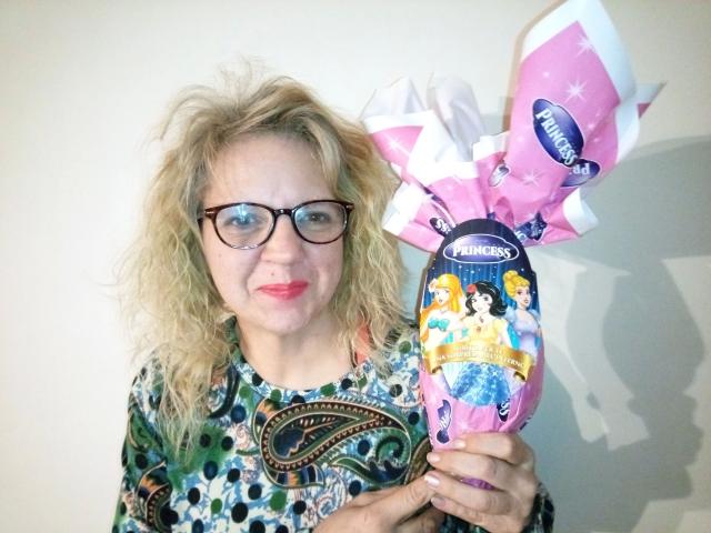 CuteDebby, carina con i capelli biondi ricci, gli occhiali e il rossetto rosso, tiene in mano un pacchetto di Twinkies rosa e bianco con un'illustrazione colorata. Indossa una camicia a righe verdi e blu..