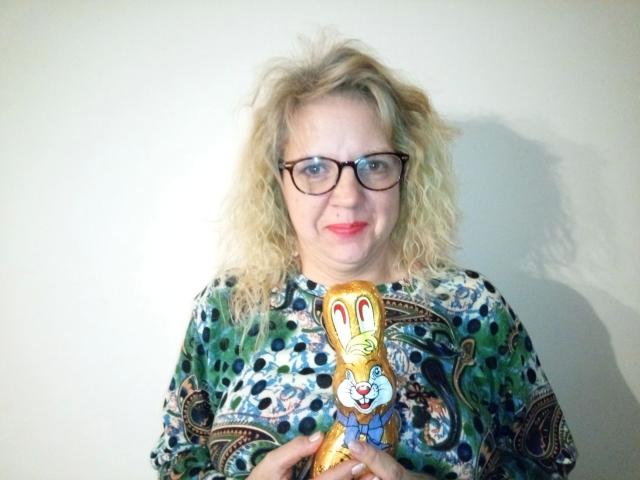 CuteDebby – hübsch mit lockigem, blondem Haar, Brille und einem bunten, gemusterten Hemd, hält eine gelbe, cartoonartige Hasenfigur mit roten Augen. Sie lächelt, sieht niedlich und ein bisschen verrückt aus..