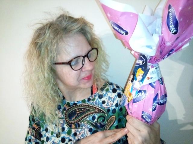 CuteDebby – attraktiv mit lockigem, blondem Haar und Brille, hält eine pinkfarbene Schachtel mit blauen und weißen Mustern, und betrachtet sie mit einem neugierigen Ausdruck..