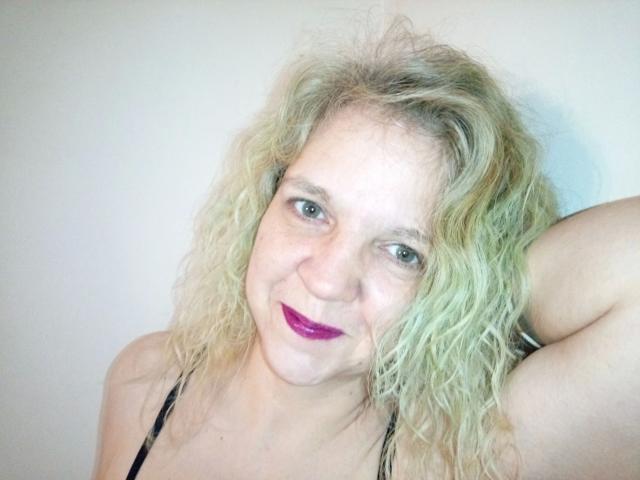 CuteDebby: bella, con capelli biondi ricci e spettinati, rossetto viola, sorridente, con un braccio alzato, vestito nero con spalline, pelle chiara, occhi azzurri, qualche lentiggine, che guarda verso la fotocamera..