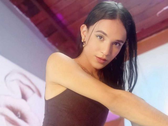 PamelaKander with long black hair, wearing a brown tank top, small gold earrings, and a gold necklace, extending her arm, looking at the camera, with a slight smile.
