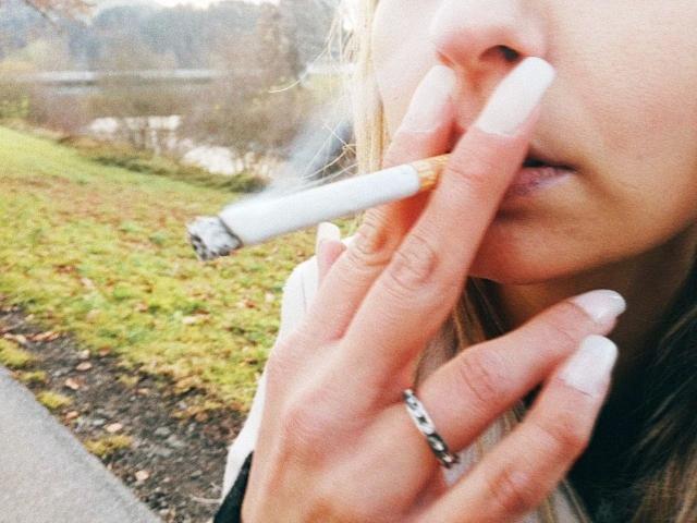 Victoria-Diamond's fingers with long, white nails holding a cigarette to her slightly open, pink-lipped mouth, wearing a silver ring, outside on a grassy path.