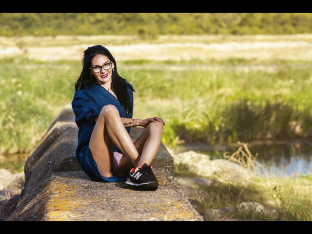SinaRauch-hot, wearing glasses, blue dress, sitting on rock, legs up, showing ass, black sneakers with white stripes, smiling, black hair, green field, sunny, relaxed.