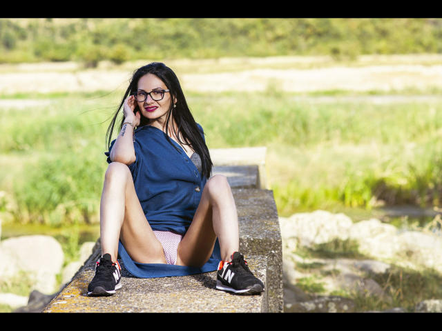 SinaRauch-hot with black glasses, blue dress, and pink panties, sitting on a stone ledge, legs spread, showing her pussy, wearing black sneakers, smiling, in a sunny outdoor setting.