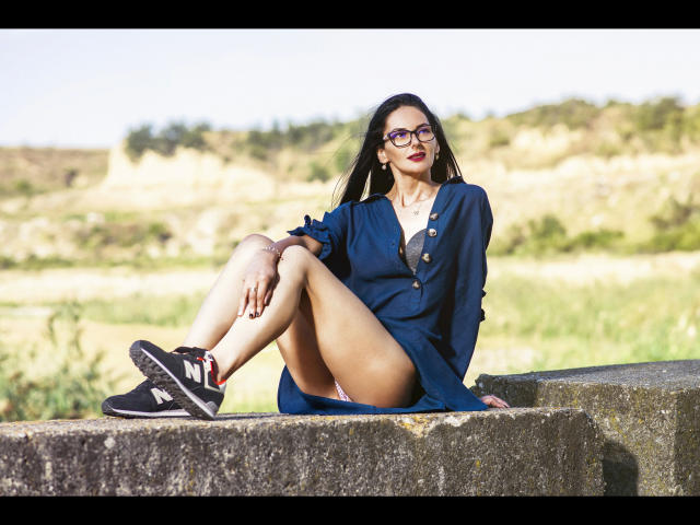 SinaRauch-hot with glasses, red lipstick, and dark blue dress, sitting on a concrete slab, legs spread, showing white panties, black sneakers with white stripes, and a sexy smile.