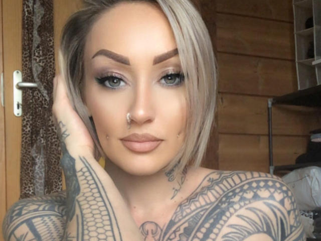 MaitresseLexia has blonde hair, tattoos, dark eyeliner, and a nose ring. Her hand touches her head, showing a tattooed arm. She has a serious expression.