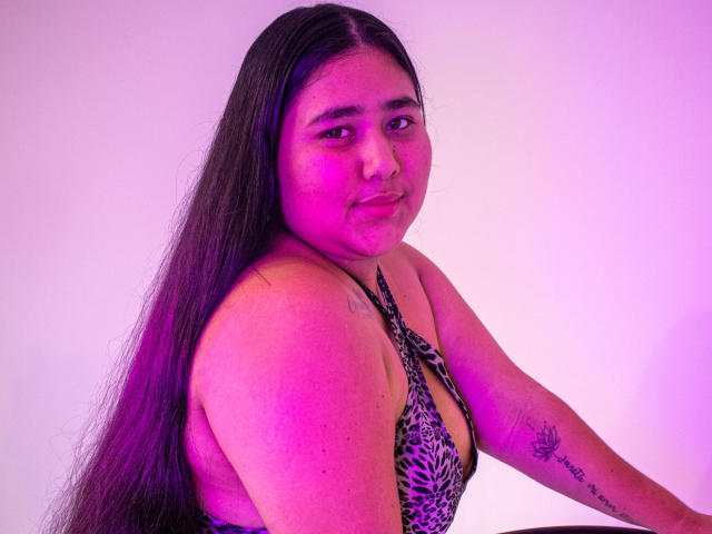 ZoeGiil with long black hair, pinkish-purple skin, leopard print top, small tattoo on right arm, looking back with a slight smile, pink-purple lighting.