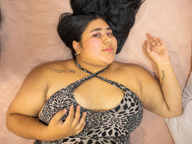 ZoeGiil with black hair lying on a pink quilt, wearing a leopard print halter top, showing large tits, tattoos on right shoulder and left wrist, smiling slightly.