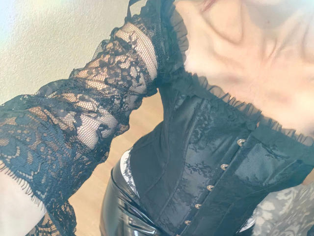 MissShine-hot wearing a black lace long-sleeve top and black lace-up corset, exposing her collarbones and part of her chest. Her skin is pale, and she has dark hair.