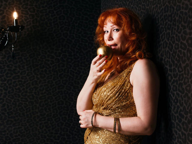 MomAimy-hot with red hair, holding a gold wine glass, wearing a shiny gold dress, standing against a dark patterned wall, looking over her shoulder, with a candle sconce.