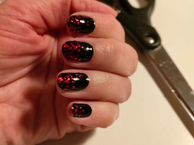 MissShine-hot's hand with black nail polish, red glitter speckles on each nail, holding scissors. Her nails are shiny, fingers slightly curled, showcasing her stylish manicure.