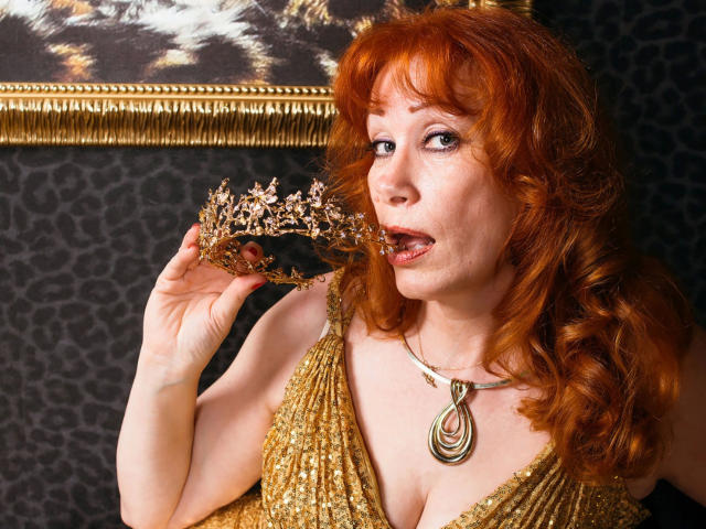 MomAimy-hot with red curly hair, gold dress, and necklace, holding a floral crown to her open mouth, looking up with a slight smile, showing her tits.