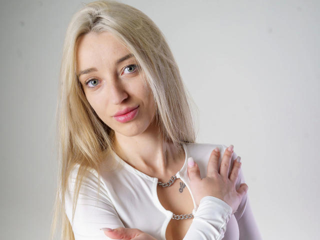 CremeDeLaCremeDE-hot has long blonde hair, blue eyes, pink lips, and a white top with a necklace. She's holding her shoulder, showing a small tattoo on her chest.