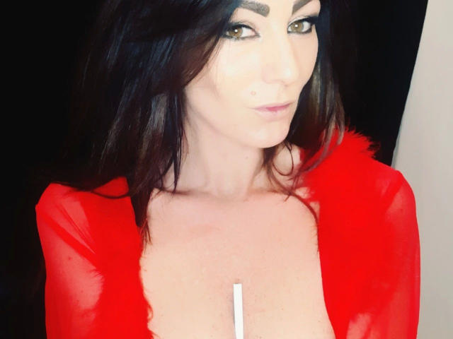 Rebella-hot with dark brown hair, wearing a red sheer top, exposing her tits, has a small white strip down the center, and a subtle smile, with green eyes.