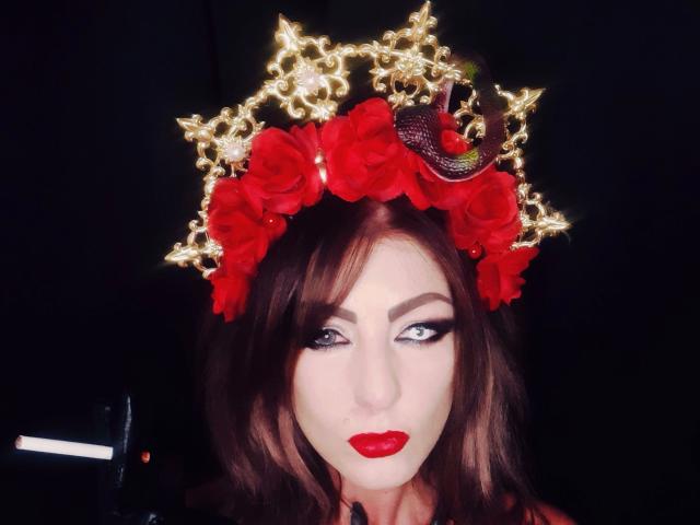 Rebella, mit roten Rosen geschmückt und einer glitzernden Krone auf dem Kopf, trägt leuchtend roten Lippenstift, Smokey-Eye-Make-up und braune, wellige Haare, vor einem dunklen Hintergrund..