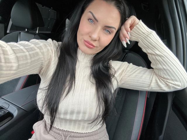 Malina-Lay, con lunghi capelli neri, occhi azzurri e labbra rosa, indossa un maglione beige a coste, con una mano tra i capelli, seduta in un'auto nera con finiture rosse..