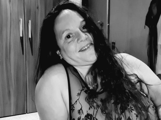 Black-and-white photo of Lady-Mercedes with long dark hair, smiling, wearing a floral-patterned top, showing her tits, standing in front of a wooden cabinet with mirrored doors.