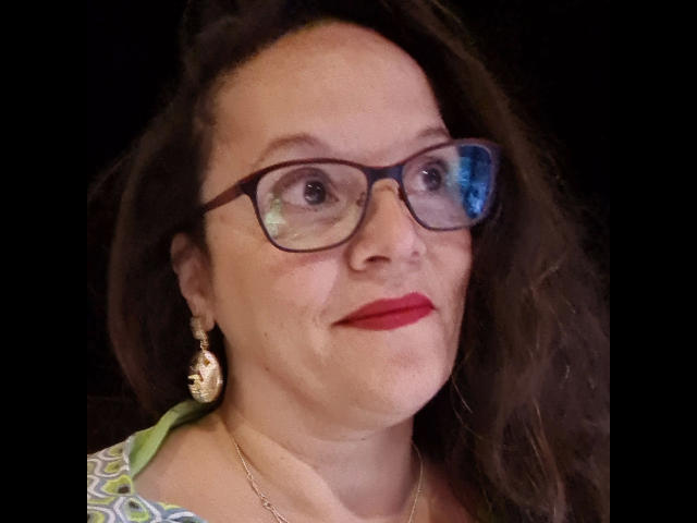 Lady-Mercedes with glasses, red lipstick, and wavy brown hair, wearing green and white patterned top, dangling gold earrings, and a gold necklace, smiling slightly.