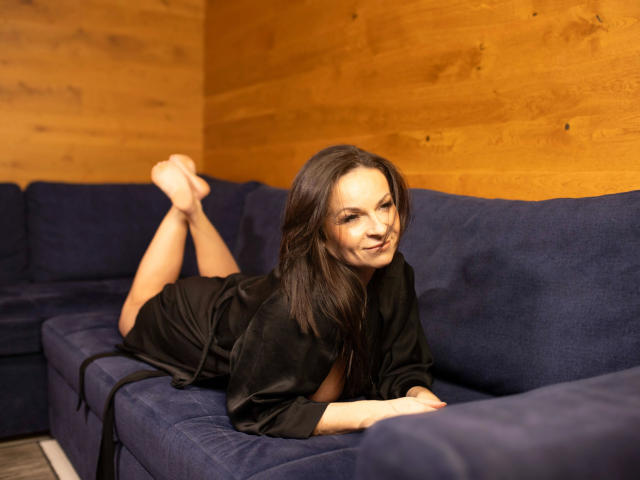 Erotic-helen, lying on a blue couch, wearing a black robe, her tits slightly visible, legs bent with feet up, wooden wall background, smiling seductively.