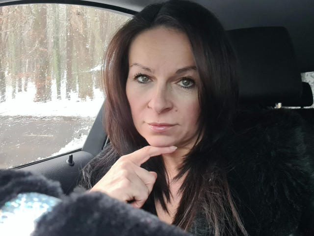 Erotic-helen with dark brown hair, green eyes, and a slight smile, wearing a black fur coat, resting her chin on her hand, inside a car. Snow outside the window.
