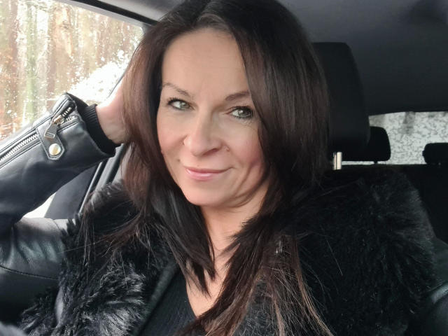 Erotic-helen with dark brown hair, green eyes, and a slight smile, wearing a black fur jacket and leather jacket, sitting in a car. Her hand rests on her head.
