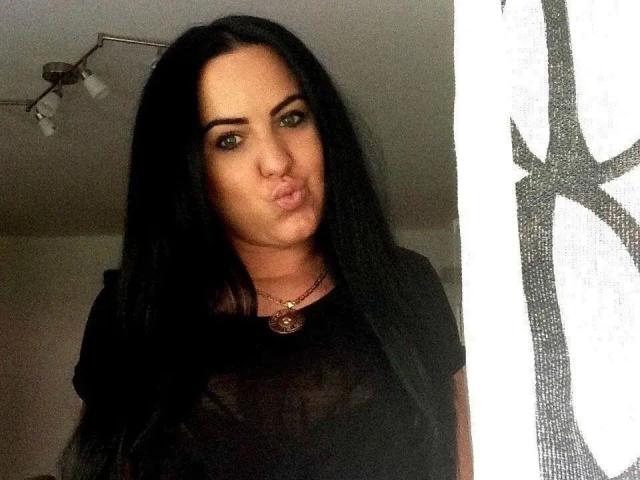 Larisssa21-hot with long black hair, pouty lips, green eyes, wearing a black top and necklace, standing near a white curtain with a black pattern.