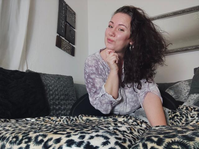 Vivien-Molinas with curly brown hair, pouty lips, wearing a gray patterned top, lying on a leopard-print blanket, resting chin on hand, looking at camera.