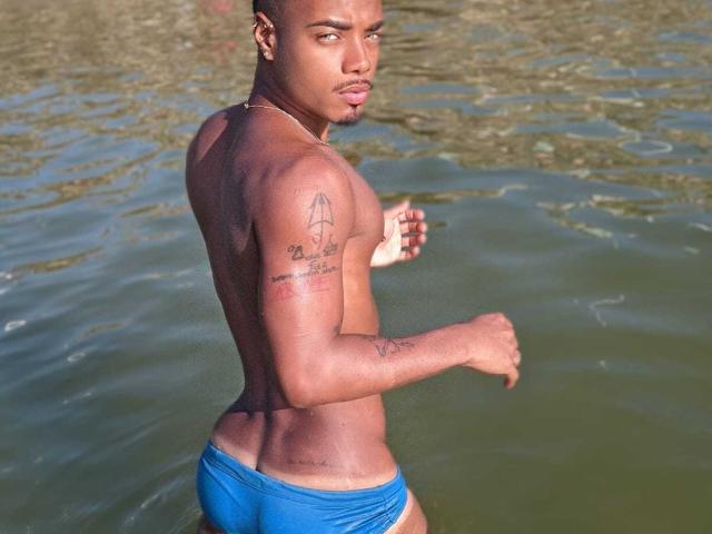 Baiano: Mit einem muskulösen, dunkelhäutigen Körper, blauen Badehosen, Tattoos am Arm und an der Schulter, blickt er ernst nach hinten und steht in flachem, grünem Wasser..