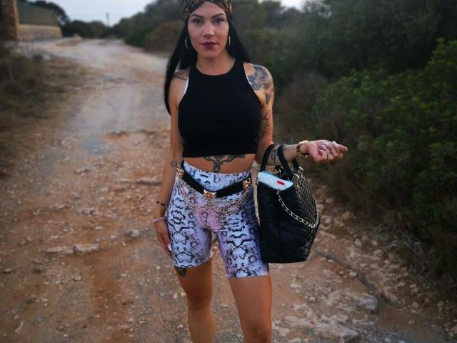 Mia124-hot with black tank top, white snake-print shorts, holding black bag, pointing, standing on dirty road, visible tattoos, long black hair, brown skin, and pink lipstick.