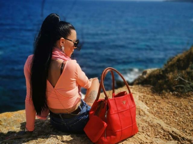 Mia124-hot with long black ponytail, pink top, denim shorts, and sunglasses, sitting on rock, red handbag, blue ocean, tattoo on left arm, sunny day.