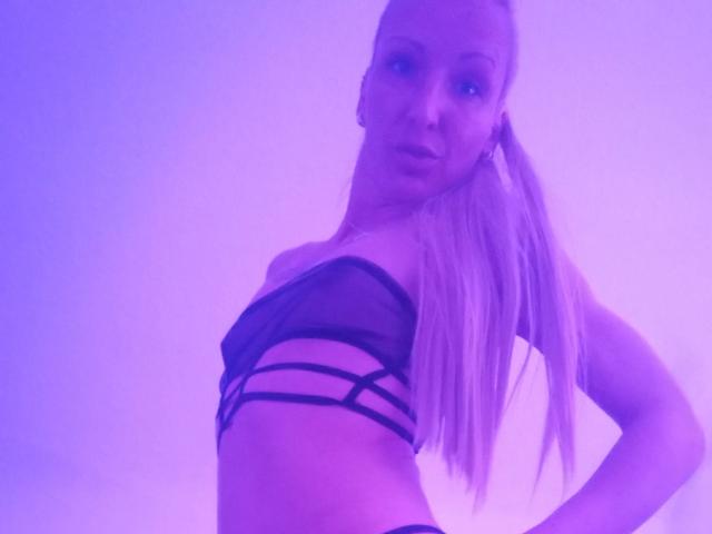 Sandyx-hot with blonde ponytail, wearing black strappy top, exposing her small tits, standing with hand on hip, bathed in purple light.
