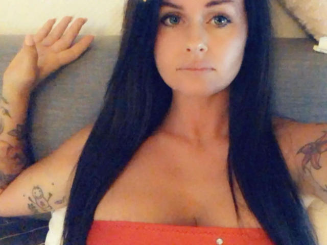CindyNight-hot with long black hair, blue eyes, and tattoos on her arms, wearing an orange strapless top, looking directly at the camera with a slight smile.