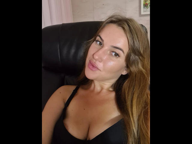 Xxjessa-hot with long brown hair, fair skin, and pink lips, wearing a black bra, sitting in a black leather chair, looking at the camera with a slight smile.