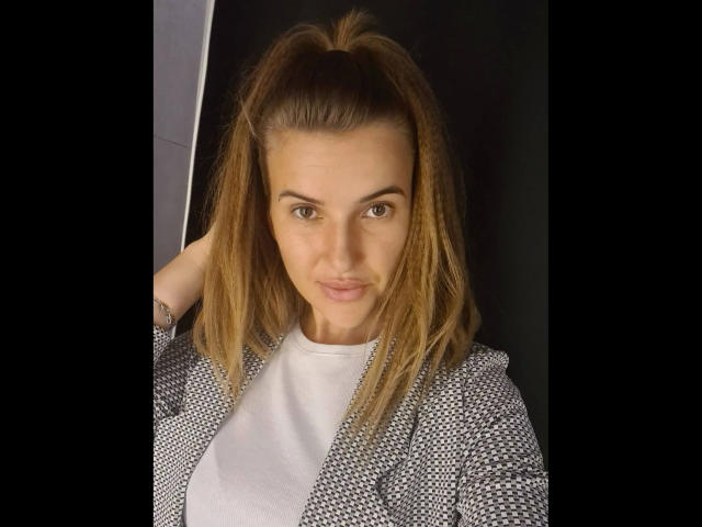 Xxjessa-hot with brown ponytail, gray patterned jacket, white tee, hand in hair, looking at camera, medium skin, brown eyes, subtle smile, no makeup, realistic selfie.
