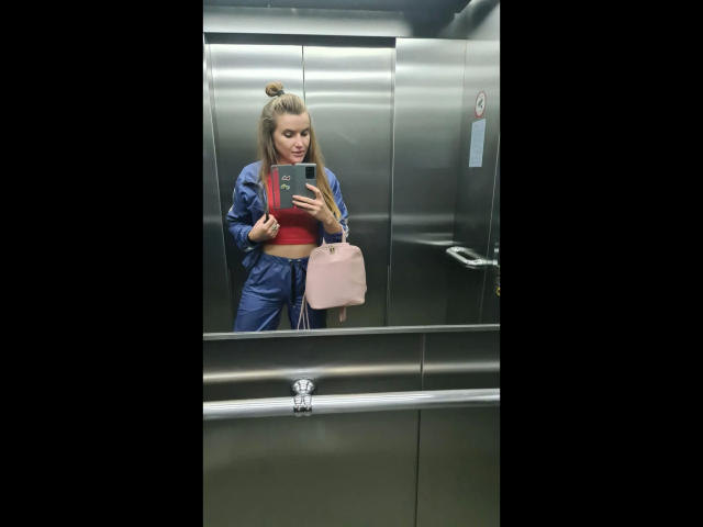 Xxjessa-hot in a elevator, wearing a blue zip-up jacket and red crop top, holding a pink purse, taking a selfie with a phone, her tits are visible.
