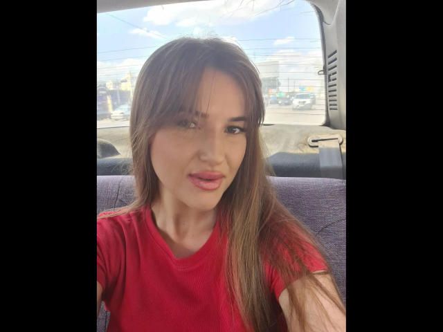 Xxjessa-hot with long brown hair, red shirt, and pink lips, sitting in a car, looking at camera with slight smile. Her ass is not visible, only her upper body.