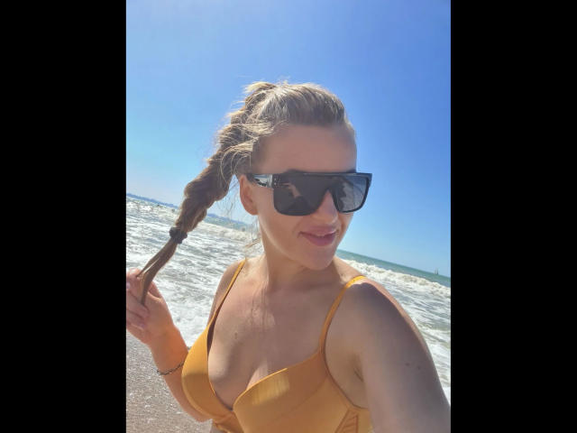 Xxjessa-hot with a braided ponytail, wearing a yellow bikini top, black sunglasses, and smiling at the camera on a sunny beach with a blue sky and ocean.