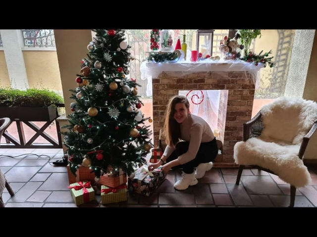 Xxjessa-hot, squatting, next to a decorated Christmas tree, wearing a white sweater, black pants, and white socks, opening a gift, in a room with a fireplace.