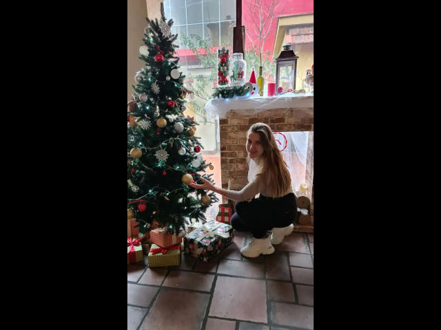 Xxjessa-hot kneeling in front of a decorated Christmas tree, touching a ornament, wearing a black top and white pants, with gifts on the tiled floor.